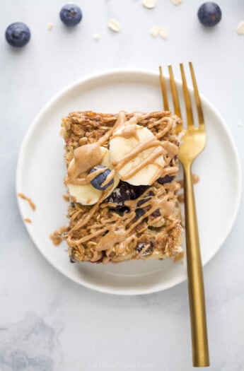 一片烤燕麦片放在白色盘子里，上面用金叉叉着，上面放着新鲜的蓝莓、香蕉片和杏仁黄油