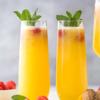 两杯桃子贝里尼酒并排放在一块灰色的切菜板上，上面有软木塞和两个新鲜的树莓